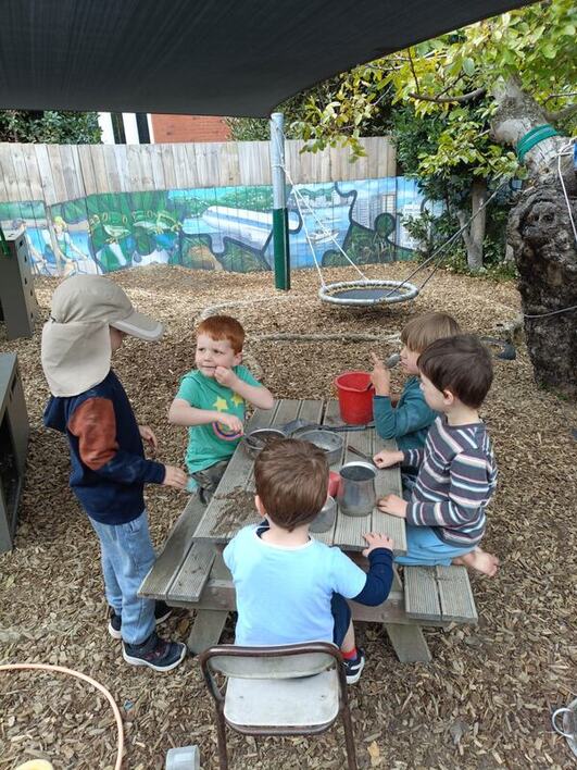 children outside at a picnic table having discussion and pretend mud and sticks lunch